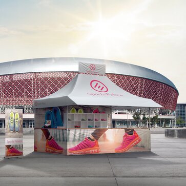 Der Faltpavillon steht vor einem Stadion und bewirbt Sportschuhe. Der Faltpavillon hat einen Banner auf dem Dach. Die Seitenwände sind bedruckt.