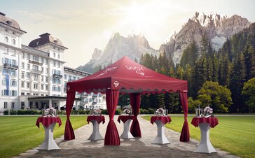 A red elegant gazebo with scalloped valance and corner curtains is located in a hotel garden. Under the gazebo are some standing tables. 