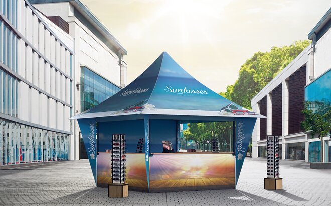 A promotion tent for sunglasses is located in the shopping street. The gazebo is blue and printed all over. 