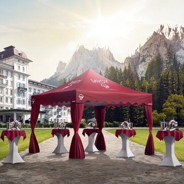 A red elegant gazebo with scalloped valance and corner curtains is located in a hotel garden. Under the gazebo are some standing tables. 
