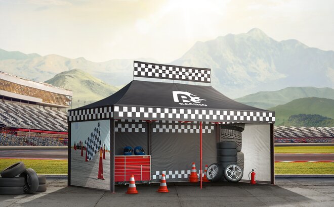 Black gazebo next to the race track. The gazebo is printed with racing patterns. Below them are tyres, cones and other motorsport accessories.