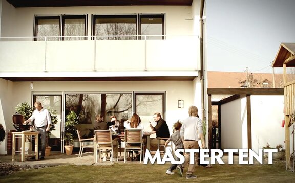 Friends at a barbecue in the garden. The man is standing at the barbecue. The guests are sitting at the table. 2 children running and playing in the garden.