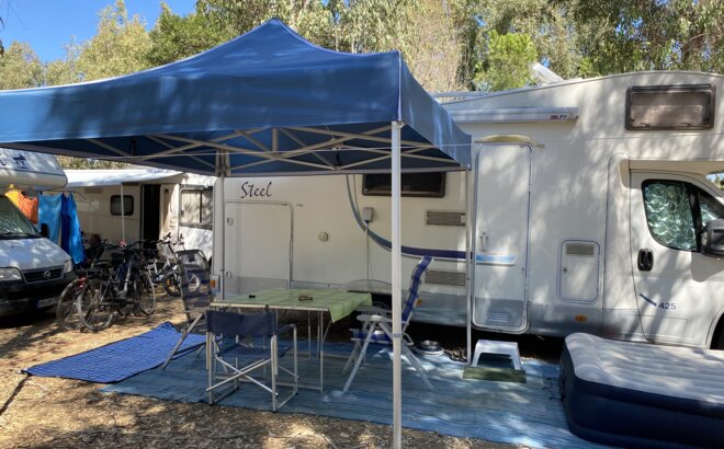 Gazebo da campeggio blu 3x3 m si trova di fronte alla roulotte del campeggio. Sotto di esso c'è un tavolo con sedie.