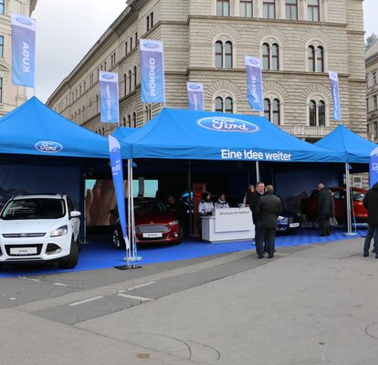 Gazebo promozionale stampato con Ford e tante bandiere blu sul tetto. Sotto il gazebo promozionale ci sono veicoli Ford.