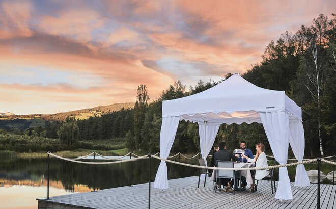 The gazebo kit Royal is located at the footbridge during sunset. Under the gazebo 4 guests are enjoying their dinner. 