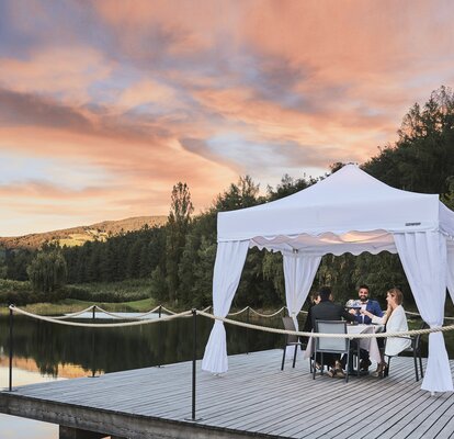 Der Faltpavillon Kit Royal steht am Steg im Sonnenuntergang. Darunter nehmen die 4 Gäste das Abendessen zu sich.