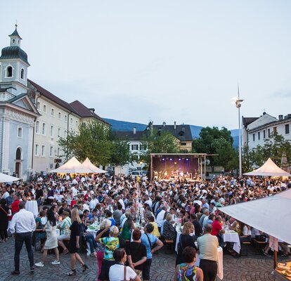 Diversi gazebo per eventi bianchi 3x3 m con tettoie nella piazza del Duomo di Bressanone al Dine & Wine festival. La gente è seduta e festeggia. Gazebo per associazioni e comuni.