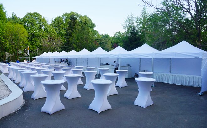 White event tents are connected to each other. In front of the gazebos are located numerous standing tables. Everything is prepared for the event. 