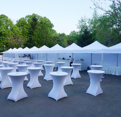 White event tents are connected to each other. In front of the gazebos are located numerous standing tables. Everything is prepared for the event. 