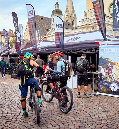 Promotion tents of BH at a bike festival on the dome square in Bressanone. The gazebo has a variety of flags. In the front are some mountain bikers. 