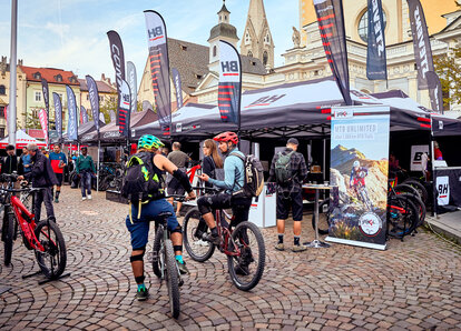 Namioty promocyjne BH podczas festiwalu rowerowego na placu przy kopule w Bressanone. W namiocie znajduje się wiele flag. Z przodu widoczni są rowerzyści górscy.