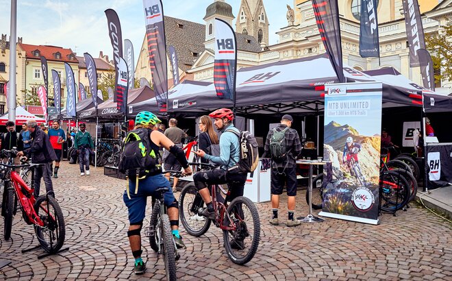 Promotion tents of BH at a bike festival on the dome square in Bressanone. The gazebo has a variety of flags. In the front are some mountain bikers. 