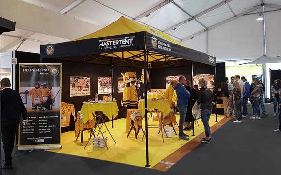 A HC Pustertal sárga vásári sátra a vásáron. Az összecsukható pavilon alatt bárasztalok és promóciós tárgyak találhatók. Emberek tolonganak a stand körül.