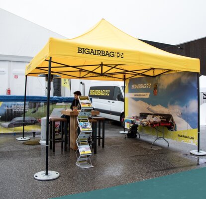 The yellow exhibition tent from Bigairbag is located at the outdoor fairground. The gazebo consists of a yellow roof with black structure. A seller is sitting below the gazebo. 