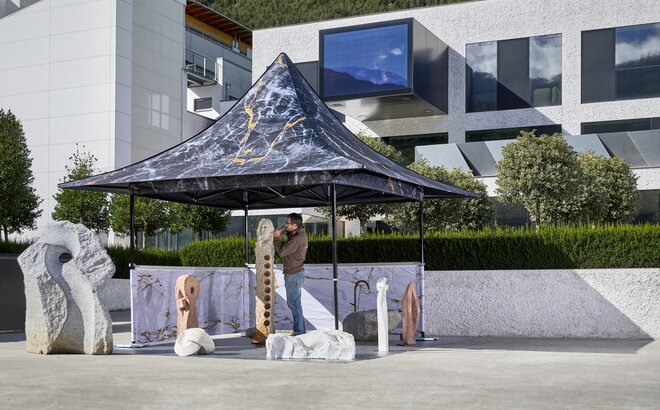The gazebo with awning is fully printed. It is located in front of a modern building and a stonemason. 