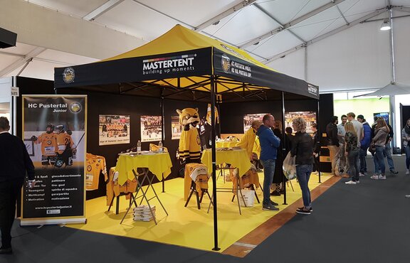 Gelbes Messezelt von HC Pustertal auf der Messe. Unter dem Faltpavillon befinden sich Stehtische und Promoartikel. Menschen tummeln sich um den Messestand.