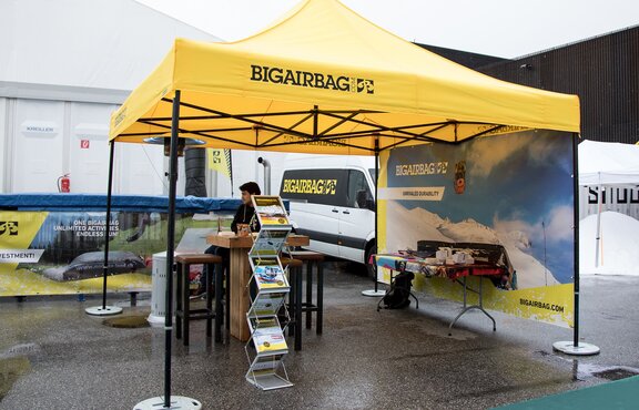 The yellow exhibition tent from Bigairbag is located at the outdoor fairground. The gazebo consists of a yellow roof with black structure. A seller is sitting below the gazebo. 