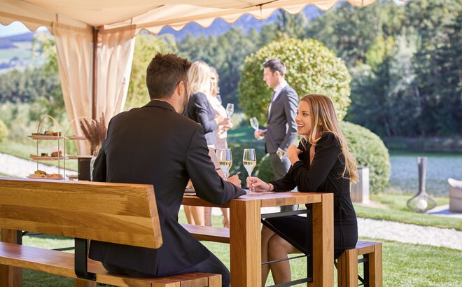 Unter einem eleganten Faltpavillon sitzt ein Paar auf einer Lago Garnitur und unterhält sich. In der Hand halten beide ein Glas Weißwein.