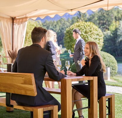 A couple is sitting under and elegant gazebo on a Lago set and is talking. Both of them are holding a glass of white wine in their hands.