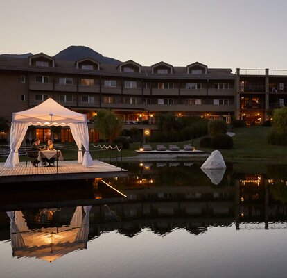 A white gastronomy tent is placed on a lighted footbridge at a lake. Underneath it a couple is having dinner. 