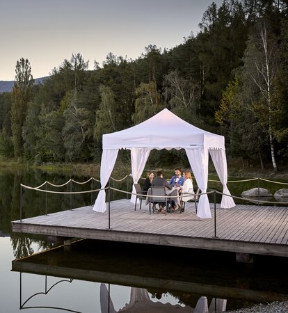 Un gazebo pieghevole per ristoranti su un molo vicino ad un lago. 4 ospiti stanno cenando sotto al gazebo bianco.