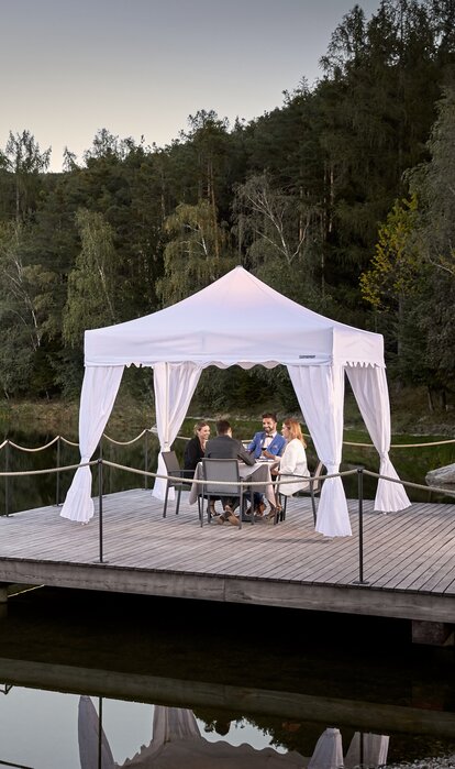 Un gazebo pieghevole per ristoranti su un molo vicino ad un lago. 4 ospiti stanno cenando sotto al gazebo bianco.