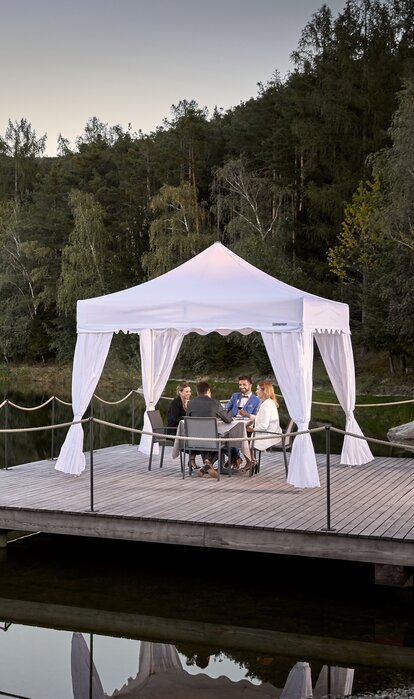 Weißer Faltpavillon steht auf dem Steg am See. Darunter dinieren 4 Personen.