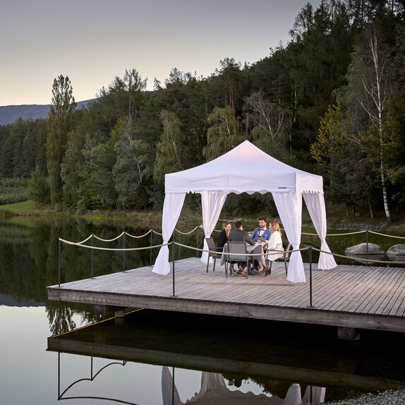 Un gazebo pieghevole per ristoranti su un molo vicino ad un lago. 4 ospiti stanno cenando sotto al gazebo bianco.