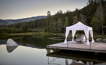 Un gazebo pieghevole per ristoranti su un molo vicino ad un lago. 4 ospiti stanno cenando sotto al gazebo bianco.
