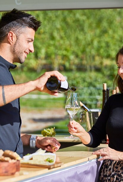 The vintner is pouring the white wine into the woman's glass. He is standing under the gazebo. She is standing in front of it.