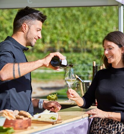 The vintner is pouring the white wine into the woman's glass. He is standing under the gazebo. She is standing in front of it.