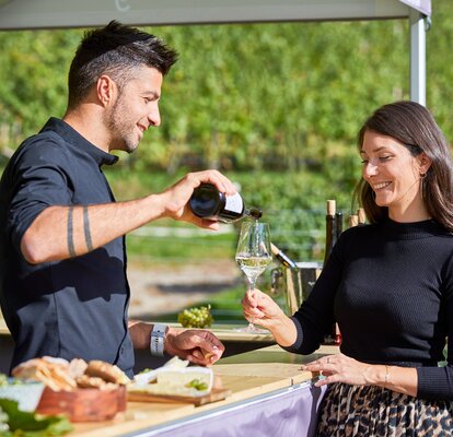 The vintner is pouring the white wine into the woman's glass. He is standing under the gazebo. She is standing in front of it.