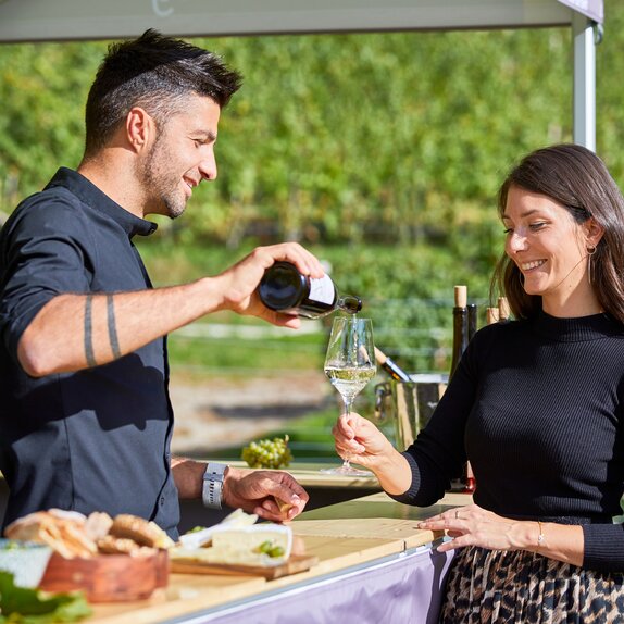 Der Winzer schenkt den Weißwein in das Glas der Frau ein. Er steht unter dem Faltpavillon und sie davor.