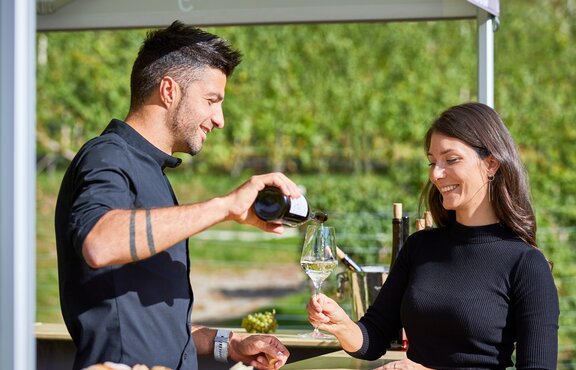 The vintner is pouring the white wine into the woman's glass. He is standing under the gazebo. She is standing in front of it.