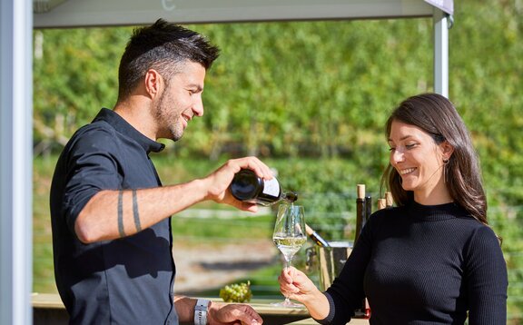 Der Winzer schenkt den Weißwein in das Glas der Frau ein. Er steht unter dem Faltpavillon und sie davor.