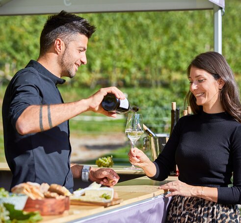 The vintner is pouring the white wine into the woman's glass. He is standing under the gazebo. She is standing in front of it.