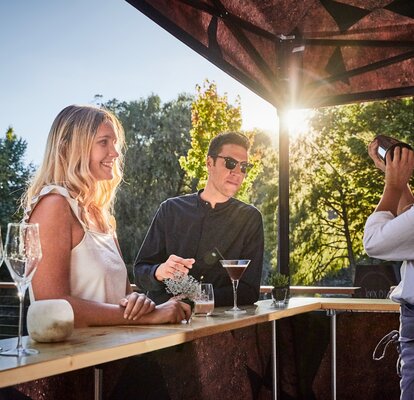 Woman is preparing a shake behind the counter for the guests in front of it. The guests are standing in front of the gazebo at sunset.