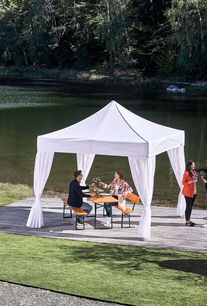 White elegant gazebo stands in the outdoor area of the restaurant on the terrace by the lake. Under the gazebo is a set of beer tables on which a couple has taken a seat. 2 people are standing next to the gazebo.
