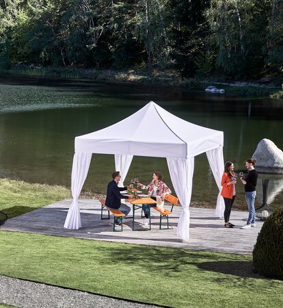 Un elegante gazebo pieghevole bianco si trova nell'area esterna del ristorante sulla terrazza in riva al lago. Sotto il gazebo pieghevole c'è un tavolo da birra apparecchiato su cui una coppia ha preso posto. 2 persone sono in piedi accanto al gazebo pieghevole.