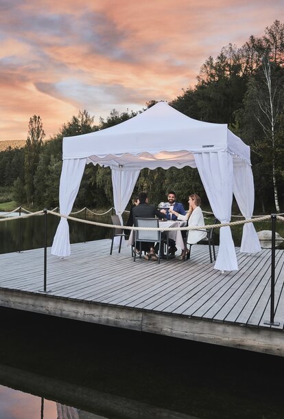 A white gazebo with corner curtains is located on the footbridge. Four people are dining underneath in the sunset. 