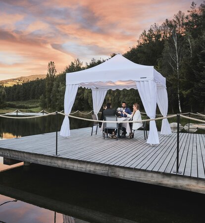 Gazebo pieghevole bianco con copripali sul molo. Le quattro presone cenano sotto il gazebo durante il tramonto. 