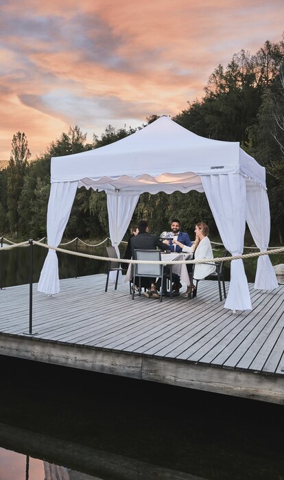 A white gazebo with corner curtains is located on the footbridge. Four people are dining underneath in the sunset. 