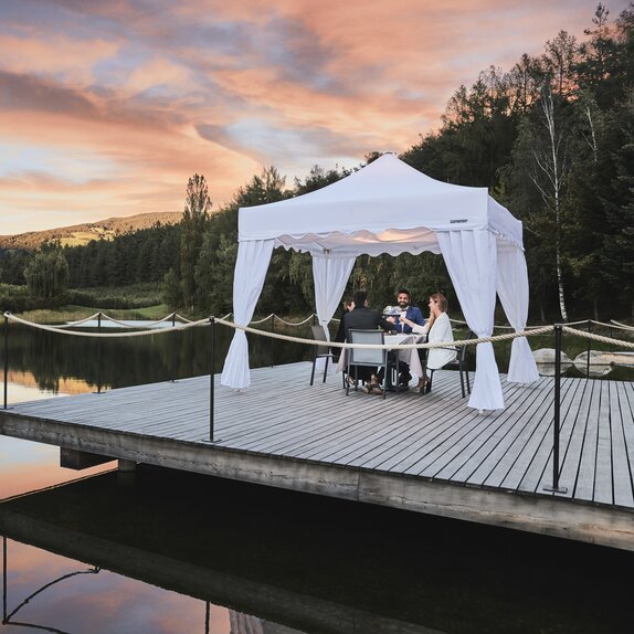 A white gazebo with corner curtains is located on the footbridge. Four people are dining underneath in the sunset. 