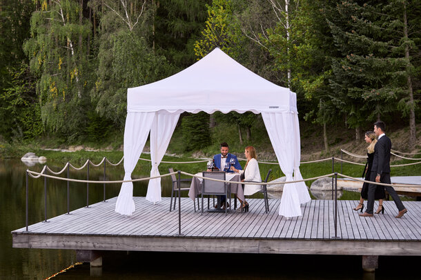 Ein Paar sitzt unter dem Faltpavillon. Das zweite Paar nähert sich. Alles ist für ein Abendessen vorbereitet.