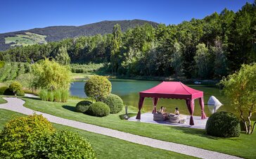 An elegant bordaux red gazebo is located directly at the lake. Underneath, a couple is sitting on two deckchairs enjoying their drinks. 