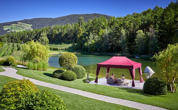 Ein eleganter bordeauxroter Faltpavillon steht direkt am See. Darunter sitzt ein Paar auf zwei Liegestühlen und genießt einen Drink.