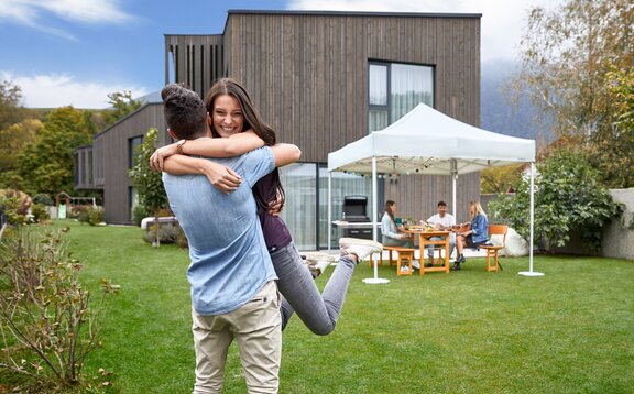 Eine Frau und ein Mann umarmen sich bei der Gartenparty. Dahinter steht der Faltpavillon, wo sich auch die anderen Gäste befinden. Im Hintergrund sieht man das Haus.