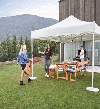 The gazebo is located in the garden. Under the gazebo a table set is prepared for a garden party. A woman with a guitar is arriving. The hostess is welcoming the woman. 