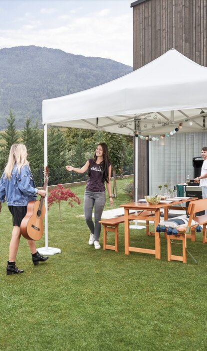 The gazebo is located in the garden. Under the gazebo a table set is prepared for a garden party. A woman with a guitar is arriving. The hostess is welcoming the woman. 