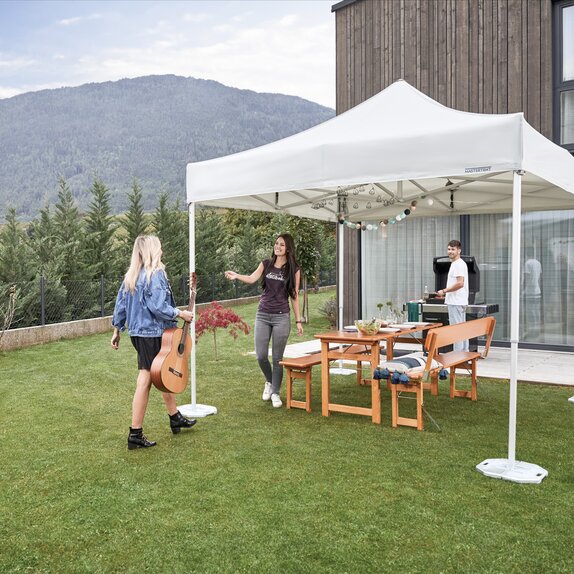 The gazebo is located in the garden. Under the gazebo a table set is prepared for a garden party. A woman with a guitar is arriving. The hostess is welcoming the woman. 